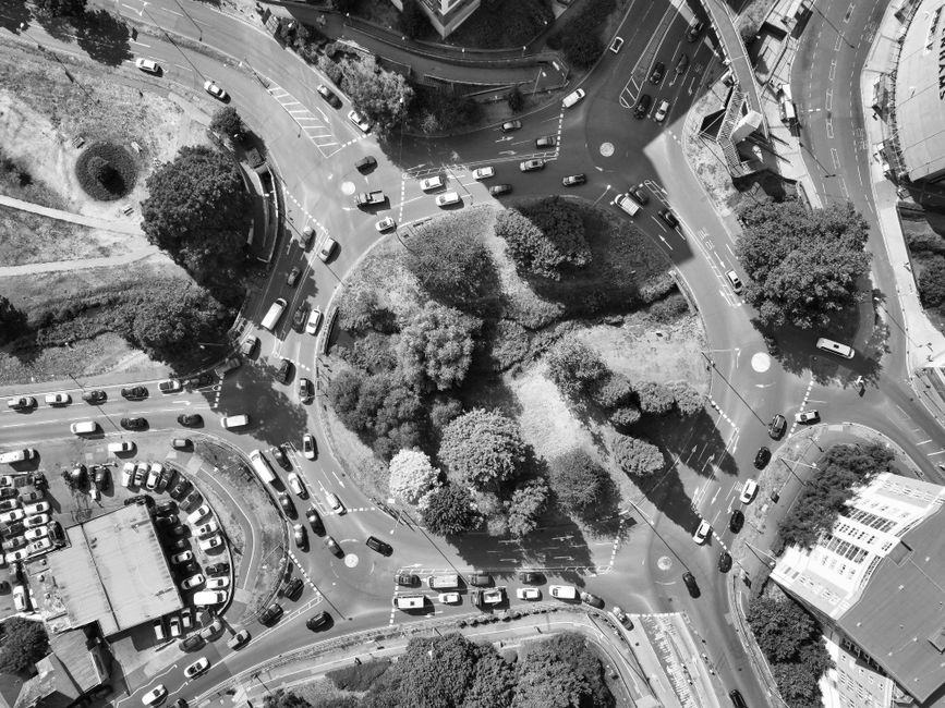 The image displays an aerial view of a busy roundabout surrounded by multiple vehicles, including cars and buses, with greenery such as trees and bushes clustered in the central island and along the r