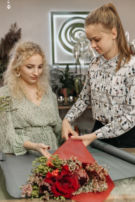 A floral arrangement being prepared indoors, featuring a bouquet with vibrant red roses, small pink and white filler flowers, and lush green foliage. The bouquet is wrapped in red floral paper, with a