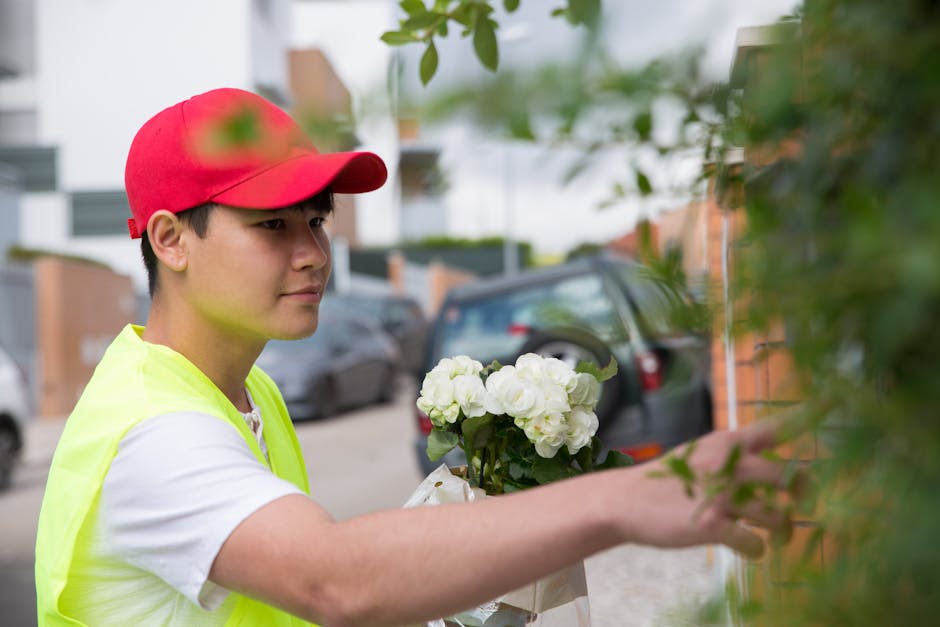 A young delivery person wearing a bright yellow vest and a red cap is placing a bouquet of fresh white roses and hydrangeas wrapped in clear cellophane and white paper against a green leafy hedge outs
