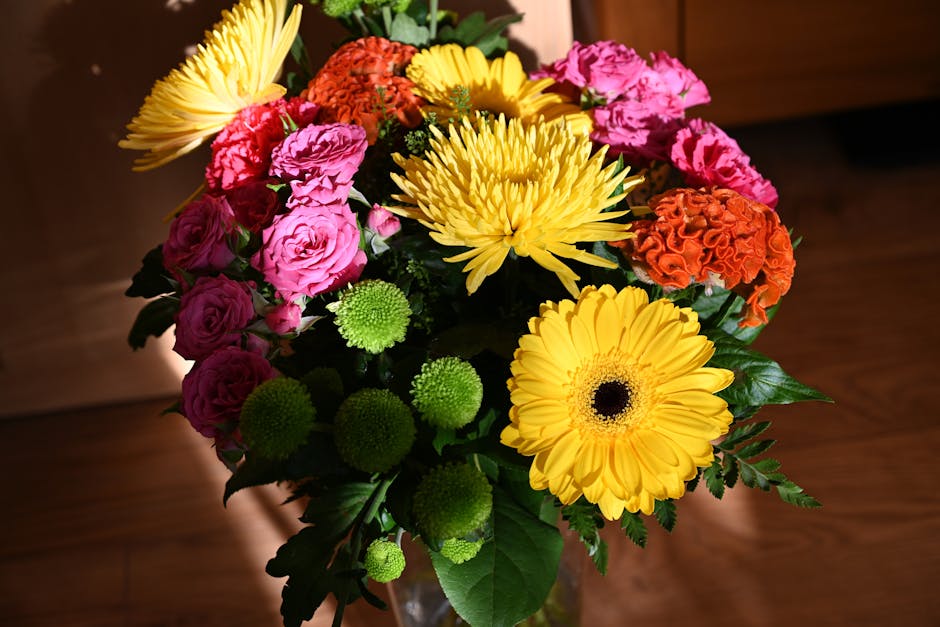 A vibrant bouquet of fresh flowers arranged in a glass vase, featuring bright yellow gerbera daisies, pink roses, orange carnations, and green chrysanthemum buds, complemented by dark green foliage. T